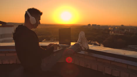 A-programmer-with-a-laptop-in-headphones-sits-on-the-roof-and-writes-code-at-sunset-and-listens-to-music.-Remote-work-freelancer.-Freedom-to-work.-Typing-on-a-keyboard-at-sunset-with-a-view-of-the-city.
